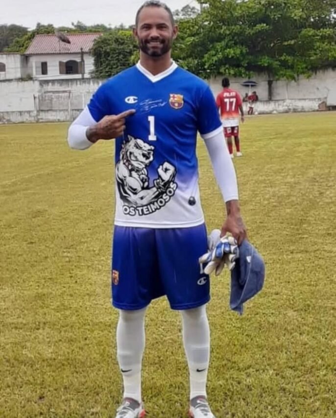 Retrato fotográfico de um goleiro, com expressão determinada. Metade de seu rosto é iluminada pelos refletores de um estádio, simbolizando fama e auge da carreira; a outra metade permanece em sombra profunda, sugerindo seu passado sombrio. Ao fundo, em desfoque, o estádio se mistura a padrões abstratos semelhantes a grades, evocando prisão.