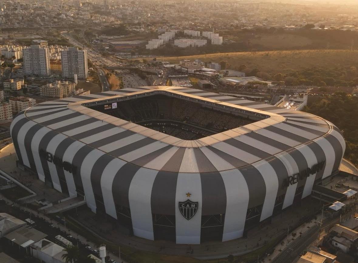 Vista aérea da Arena MRV ao pôr do sol, cobertura listrada em cinza e branco e a cidade ao fundo com luz dourada.