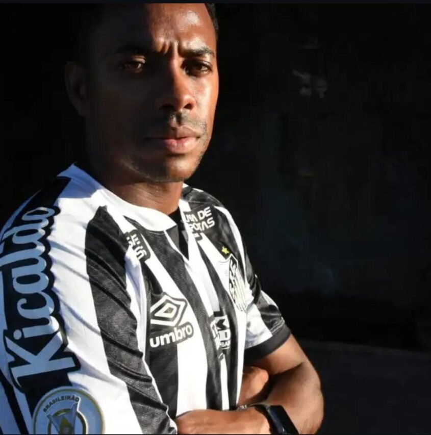 Jogador com camisa listrada preta e branca, braços cruzados, meia-luz lateral destacando expressão séria; fundo escuro.