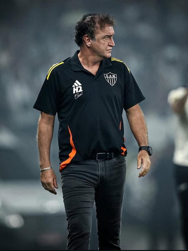 Treinador do Atlético Mineiro à beira do campo, camisa preta com detalhes laranja e ombros amarelos, olhar sério; fundo de estádio desfocado.