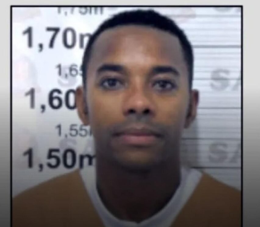 Retrato frontal de um homem diante de painel claro com marcações de altura em metros; expressão neutra, olhar direto à câmera.