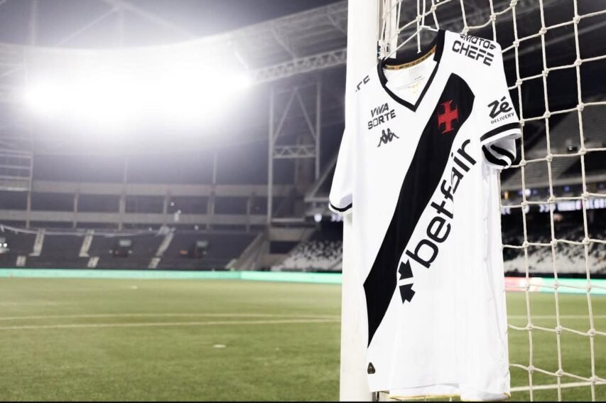 Camisa branca do Vasco da Gama pendurada na rede do gol em um estádio iluminado à noite.