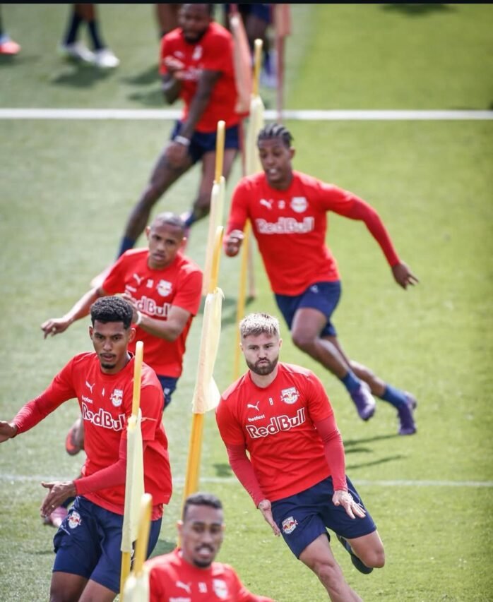 ogadores de futebol com camisas vermelhas “Red Bull” fazem exercício de mudança de direção entre estacas amarelas em um campo gramado.