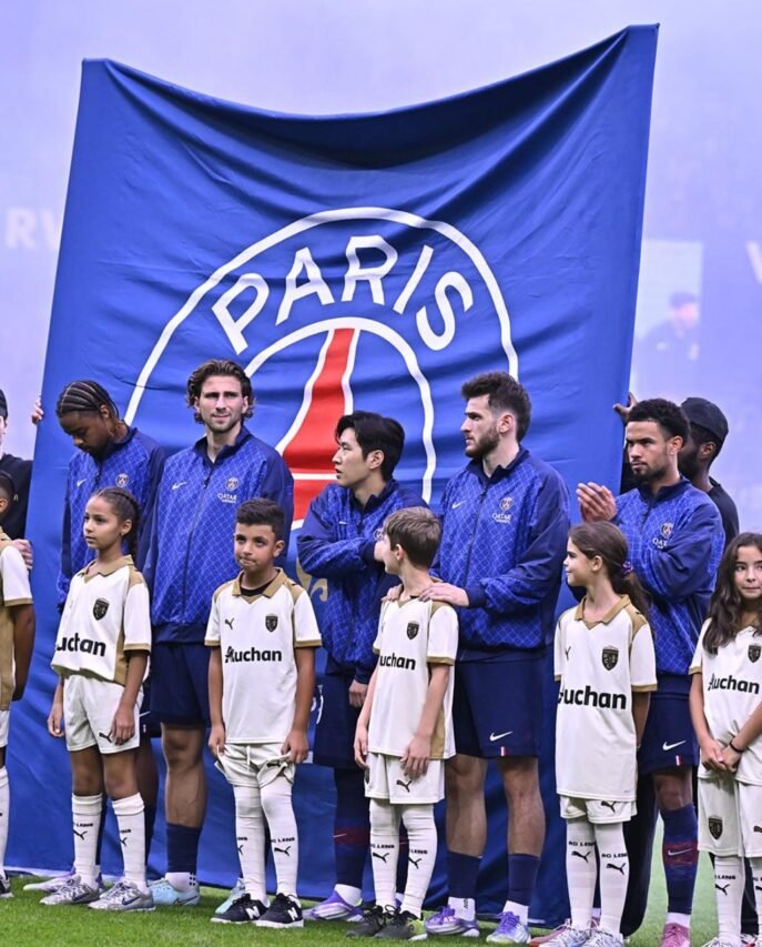 Jogadores do Paris Saint-Germain, de agasalhos azuis, alinham-se ao lado de crianças mascote com uniformes branco e dourado diante de um bandeirão azul do PSG com a palavra “PARIS” e a silhueta da Torre Eiffel.