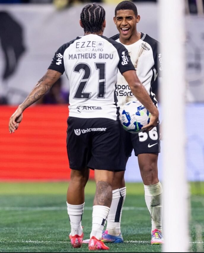 Dois jogadores de futebol com uniforme preto e branco se aproximam no gramado; o camisa 21, de costas, abre os braços enquanto o companheiro nº 56 sorri segurando a bola.