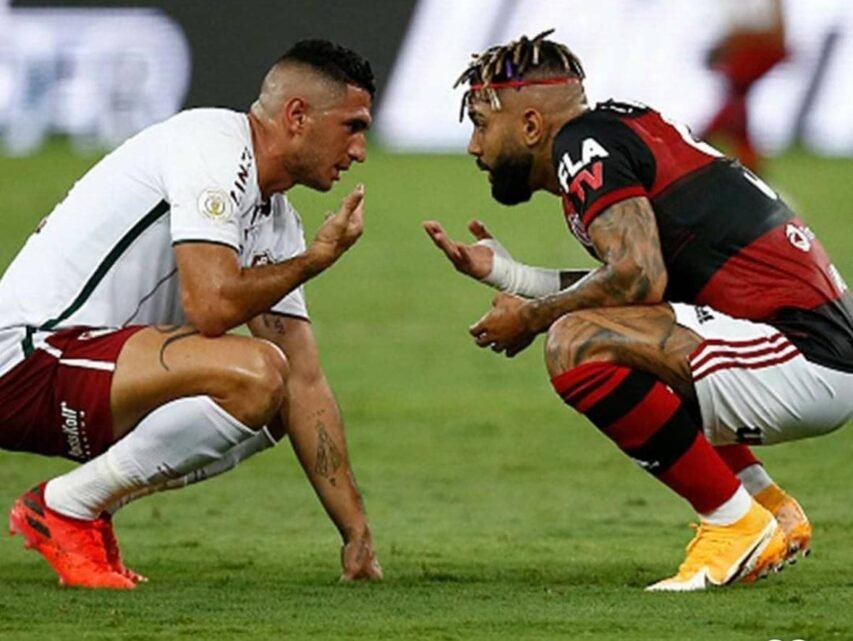 Dois jogadores, um do Fluminense e outro do Flamengo, agachados frente a frente em campo, gesticulando intensamente, em um momento de tensão ou provocação durante um clássico.