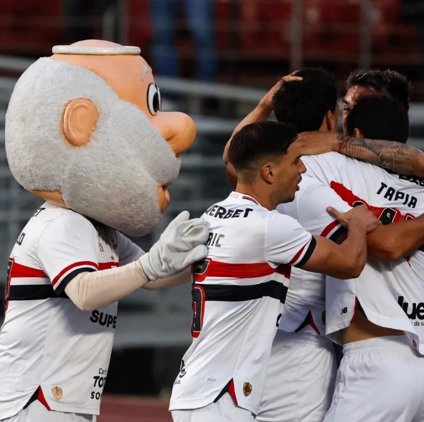 Mascote de barba branca do São Paulo Futebol Clube comemora junto aos jogadores que se abraçam após um gol.
