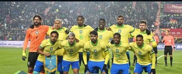 Onze jogadores do Brasil posam para a foto oficial antes do jogo, com faixa verde à frente escrita “ELIMINATORIAS SUDAMERICANAS COPA DEL MUNDO FIFA 2026”; estádio lotado e noite fria ao fundo.