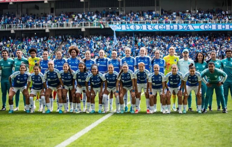 Elenco completo do Cruzeiro feminino posando para foto oficial antes de partida, com a torcida celeste ao fundo.
