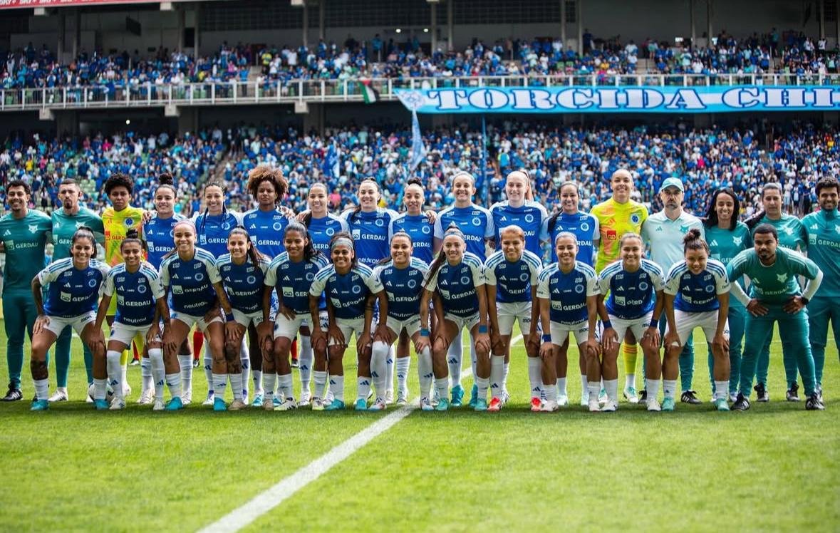 Elenco completo do Cruzeiro feminino posando para foto oficial antes de partida, com a torcida celeste ao fundo.