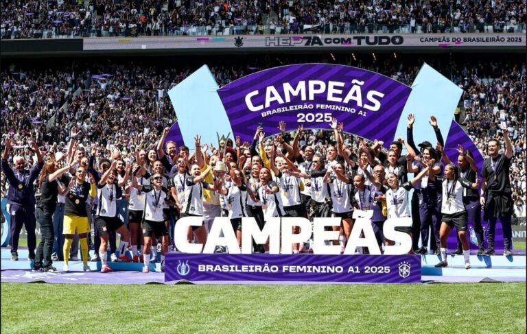 Elenco do Corinthians celebra no pódio com arco roxo “CAMPEÃS 2025” e faixa “Brasileirão Feminino A1 2025”; jogadoras com medalhas erguem a taça; arquibancadas lotadas ao fundo.