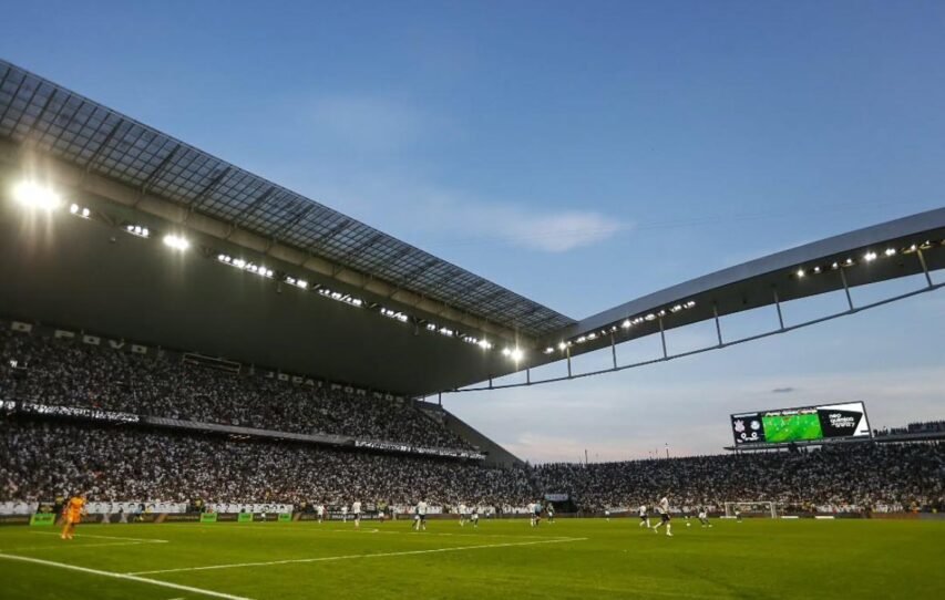 Arena lotada com torcedores acompanhando partida de futebol à noite sob luzes de refletores.