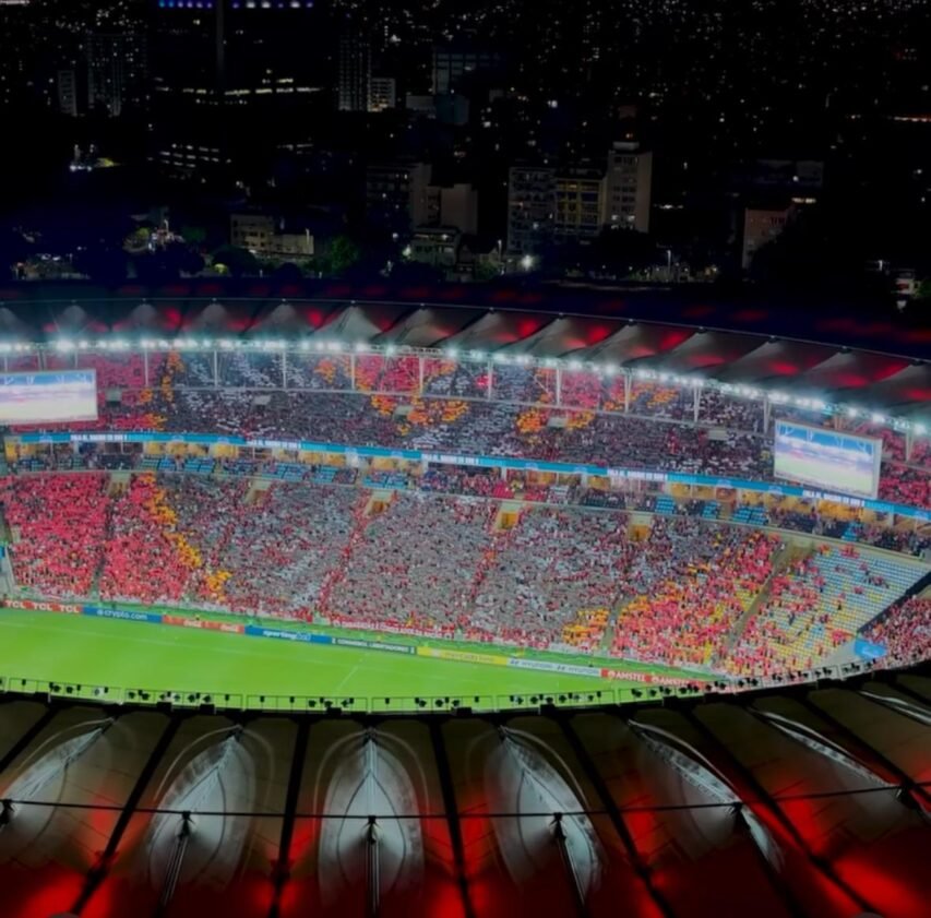 Vista interna do Estádio do Maracanã durante a noite, com as arquibancadas lotadas de torcedores em vermelho, amarelo e cinza. As luzes refletem no gramado verde e iluminam intensamente a atmosfera do jogo.