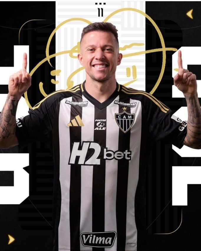 Jogador do Atlético-MG sorridente, vestindo o uniforme alvinegro, comemorando com os braços erguidos e dedos apontando para cima.